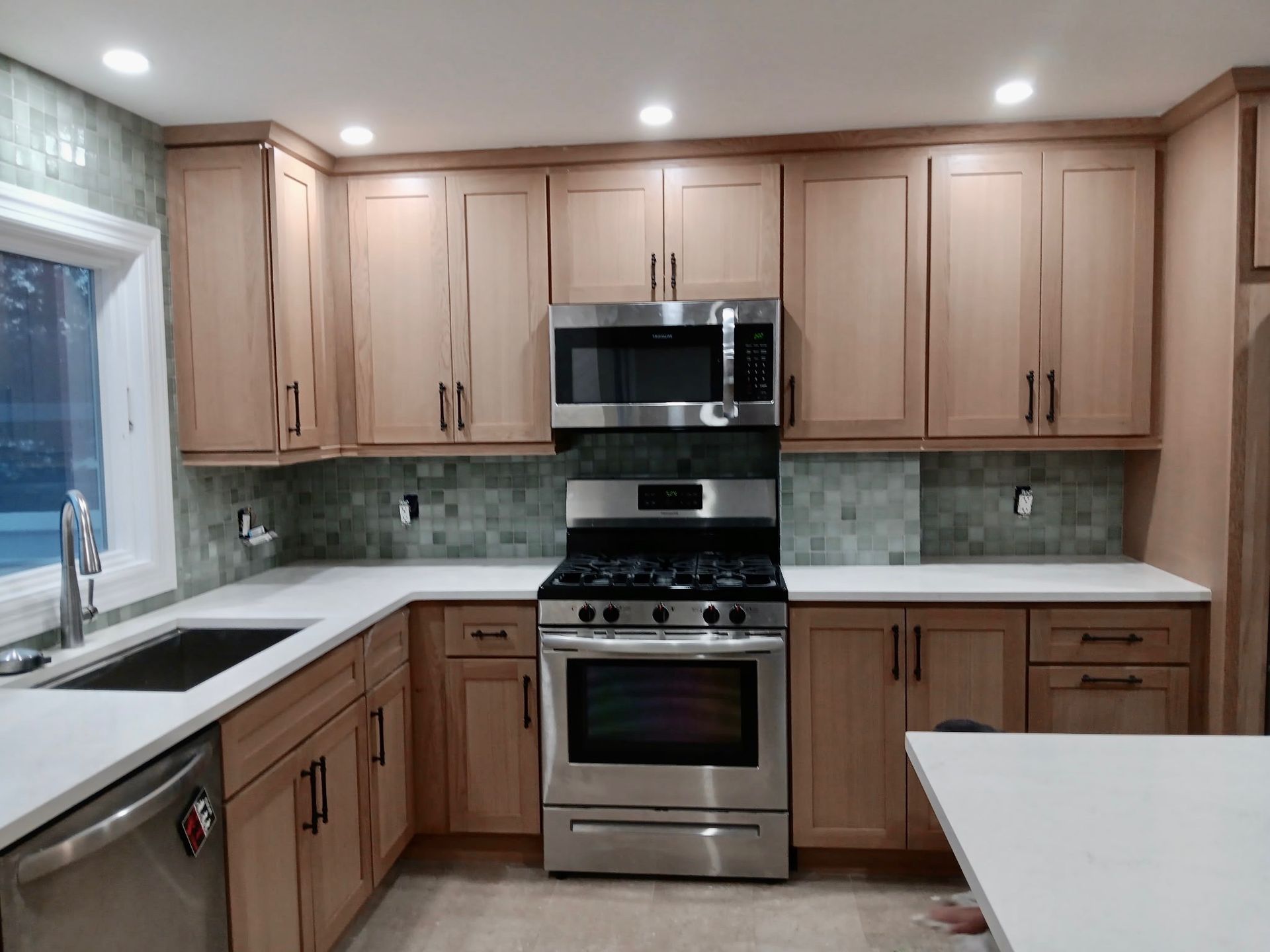Kitchen with light wood cabinets, stainless steel appliances, white countertops, and green backsplash.