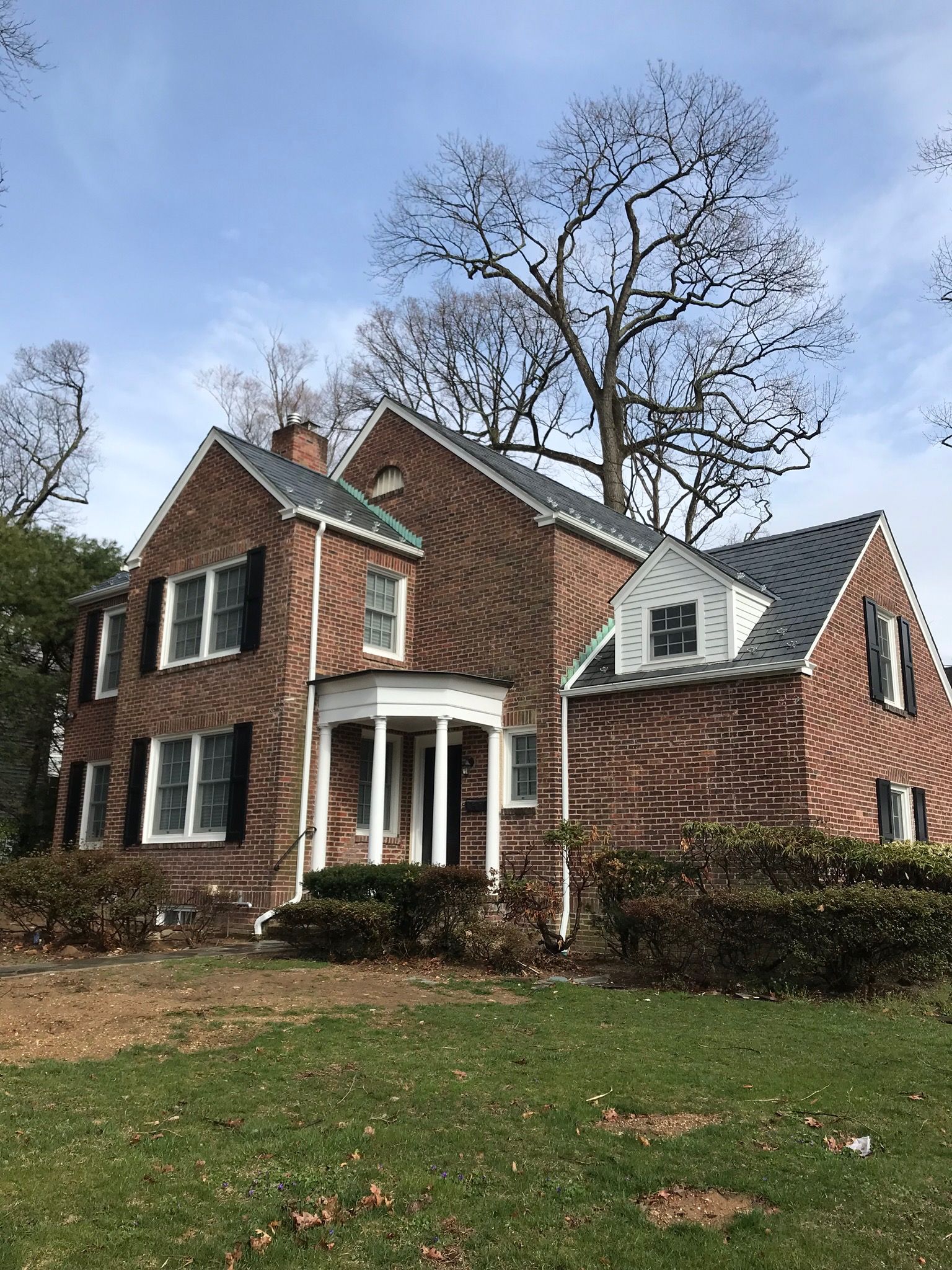 Brick house with dark shutters, white trim, and a small porch, set on a green lawn with a large tree in the background.