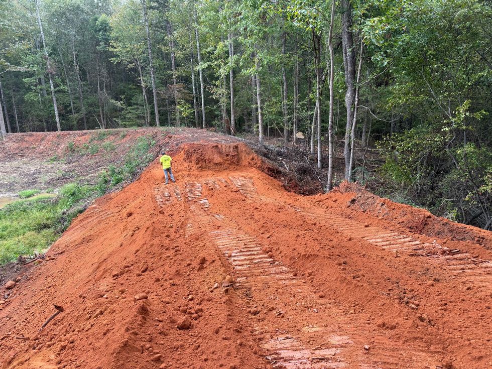 Freshly graded red dirt road through a forest, with a worker in a yellow shirt in the distance.