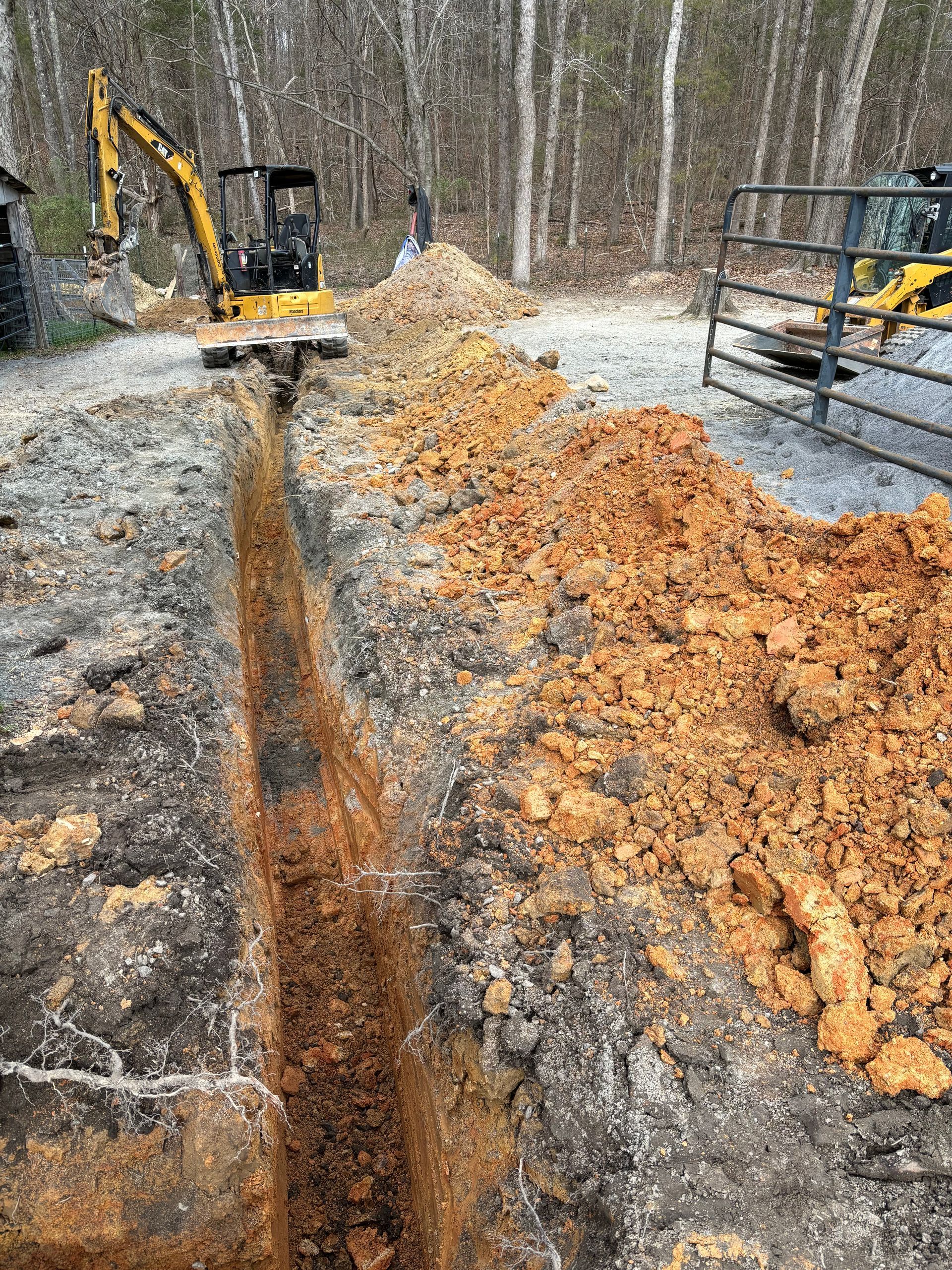 Trench being dug in a wooded area by an excavator, with piles of orange dirt beside it