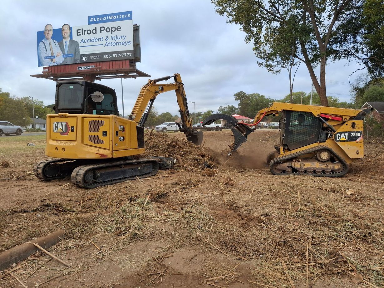 Yellow excavator demolishing a small yellow machine in a dirt lot beside a billboard