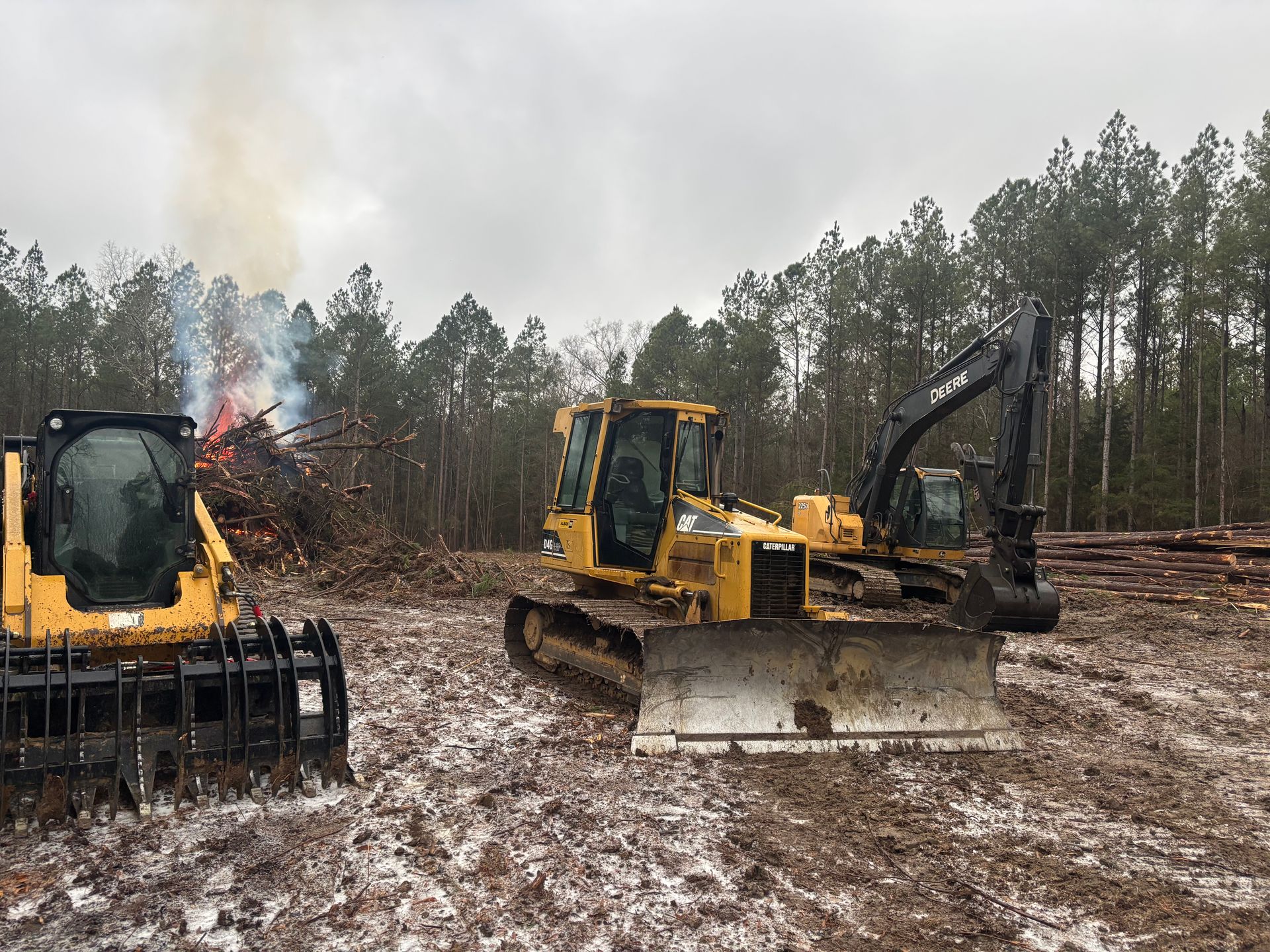 Yellow bulldozers on a muddy, snowy worksite with smoke rising in a forest clearing