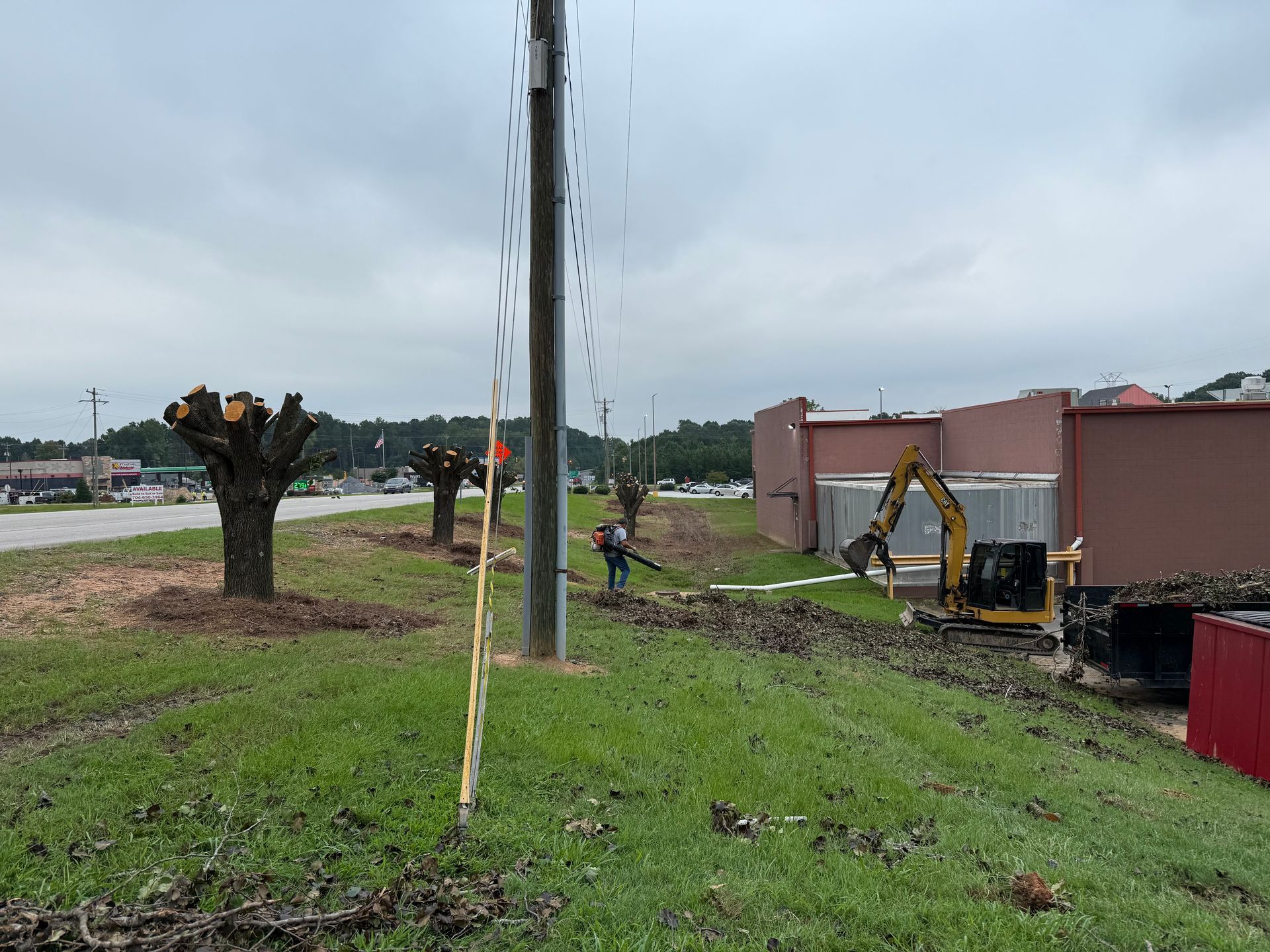 Construction equipment working beside a grassy roadside near a pink building and utility pole