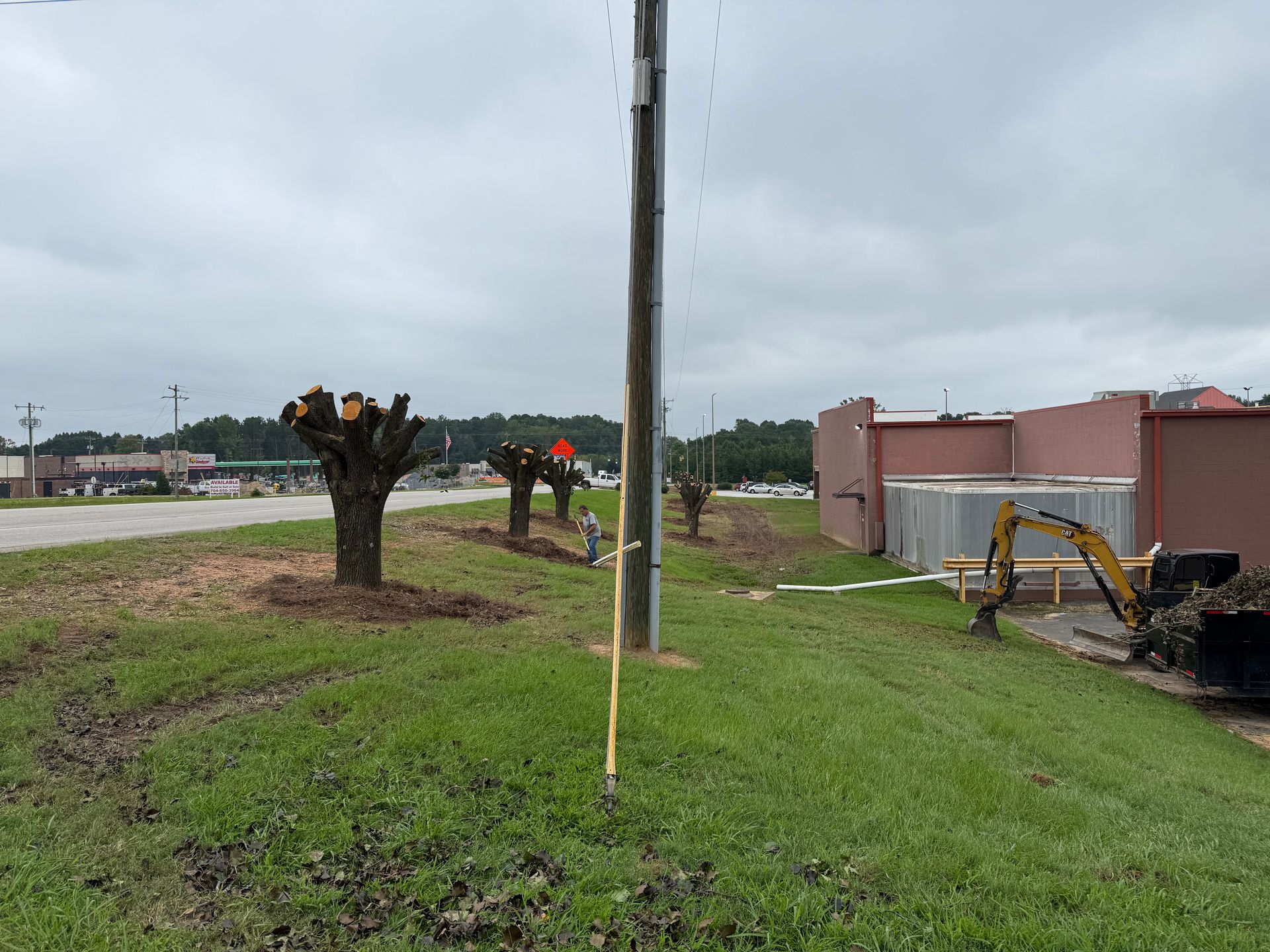 Storm-damaged roadside with trimmed trees, utility pole, and construction equipment beside a pink building