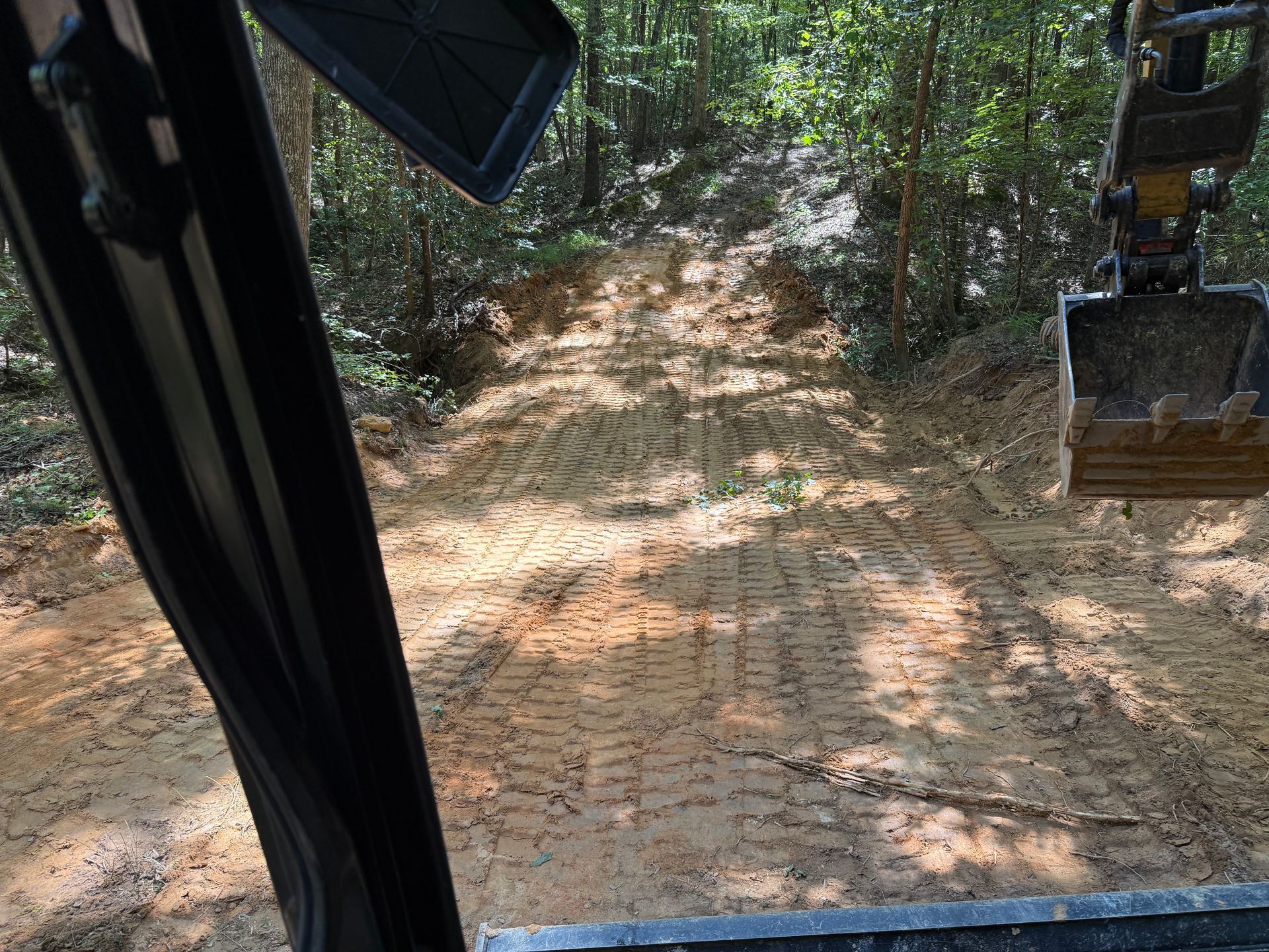 View from a construction vehicle of a dirt trail in the woods with excavator bucket visible on the right