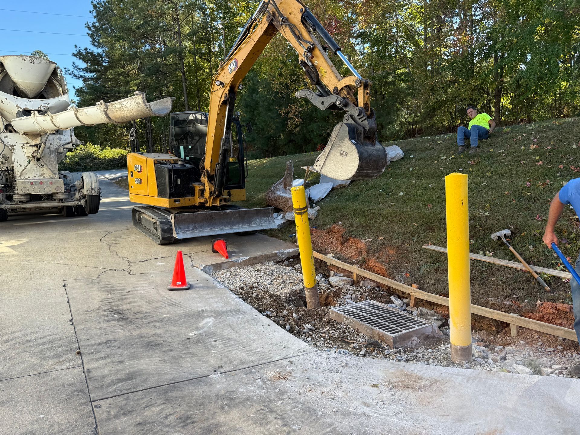 Yellow excavator digs beside a sidewalk with orange cones and workers near a fallen tree trunk.
