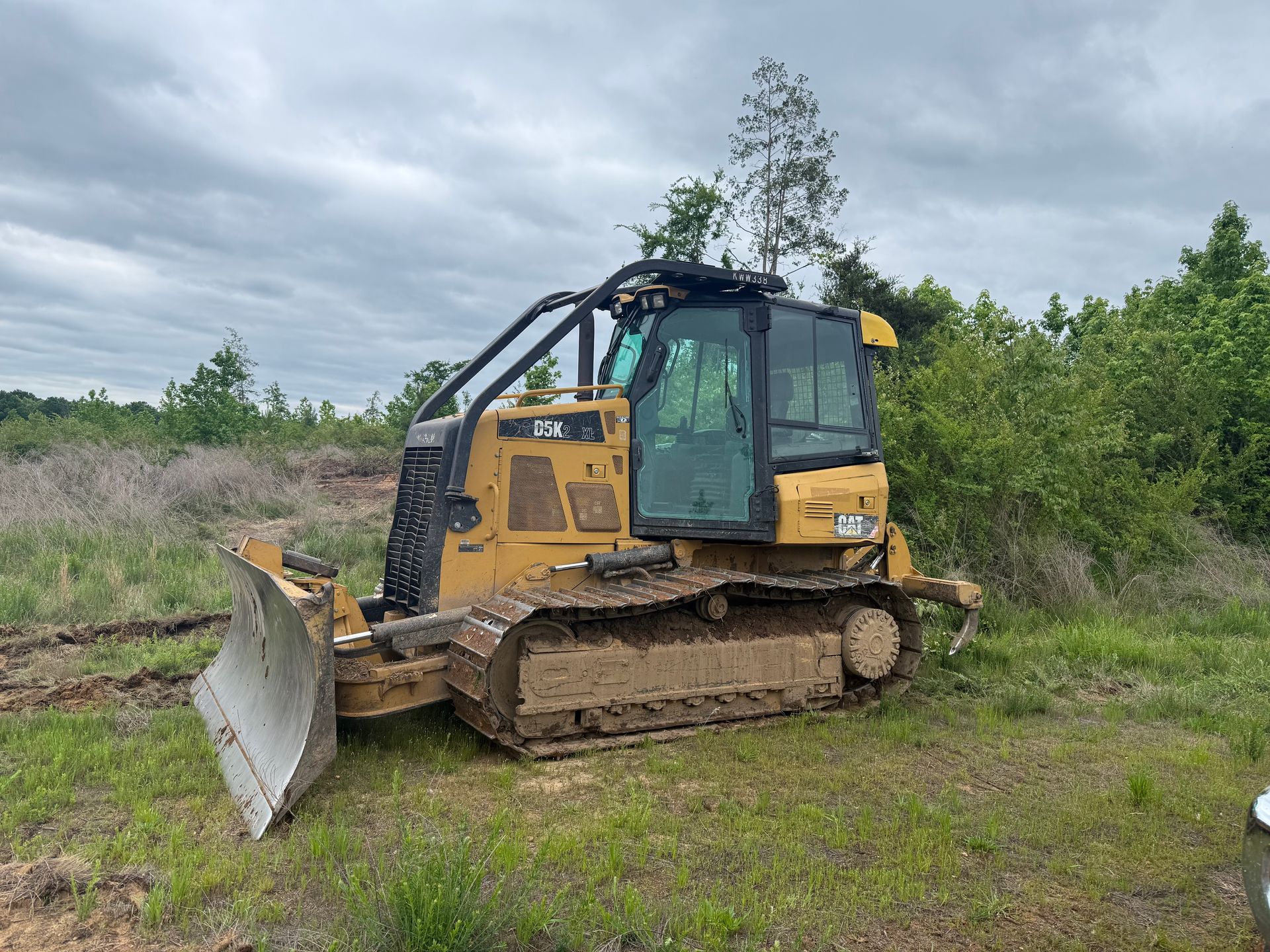 Yellow bulldozer with front blade parked on muddy ground beside trees under a cloudy sky