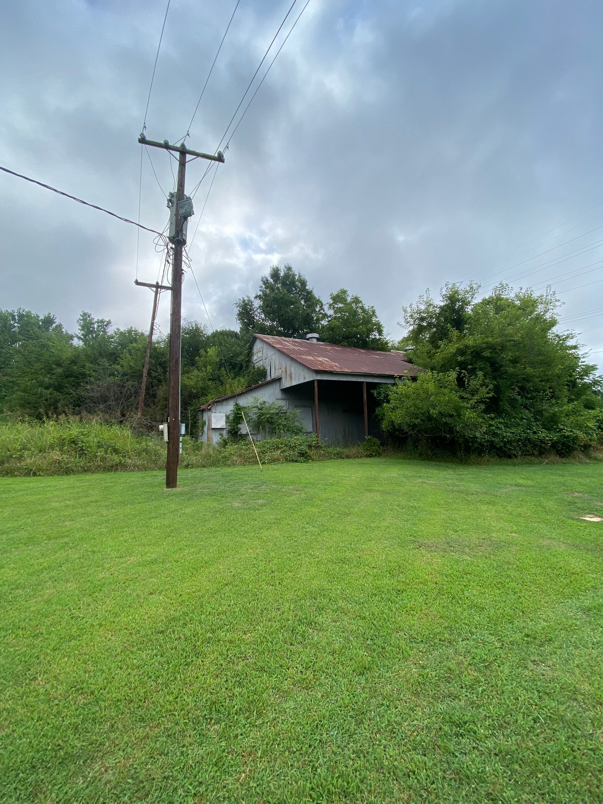 Small house with a rusty roof beside a utility pole in a lush green field under cloudy sky