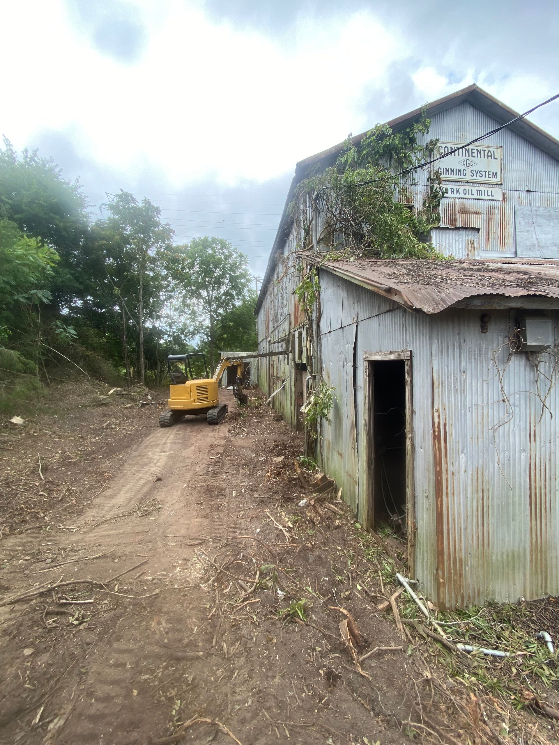 Dirt road beside a weathered corrugated metal shed, with a small yellow excavator near trees and cloudy sky