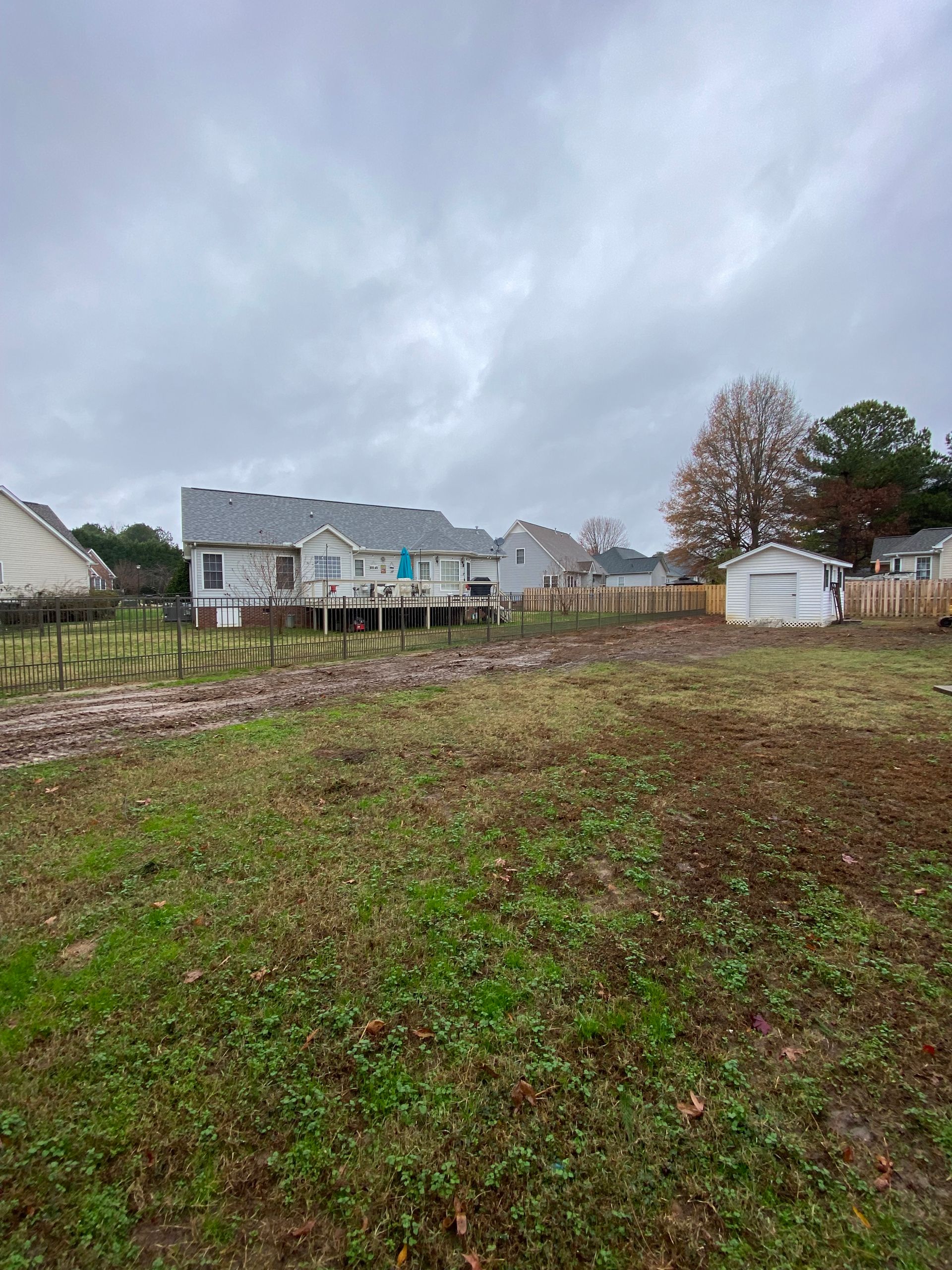Overcast backyard with muddy grass, fenced yard, and houses in the distance