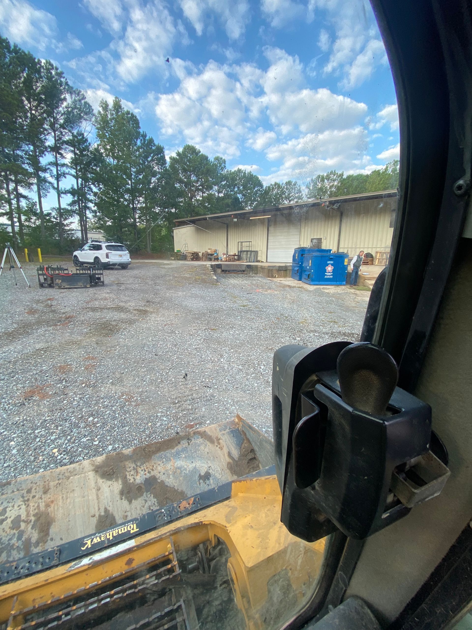 View from inside a construction vehicle of a gravel lot with a white truck, blue dumpster, and trees by a building.