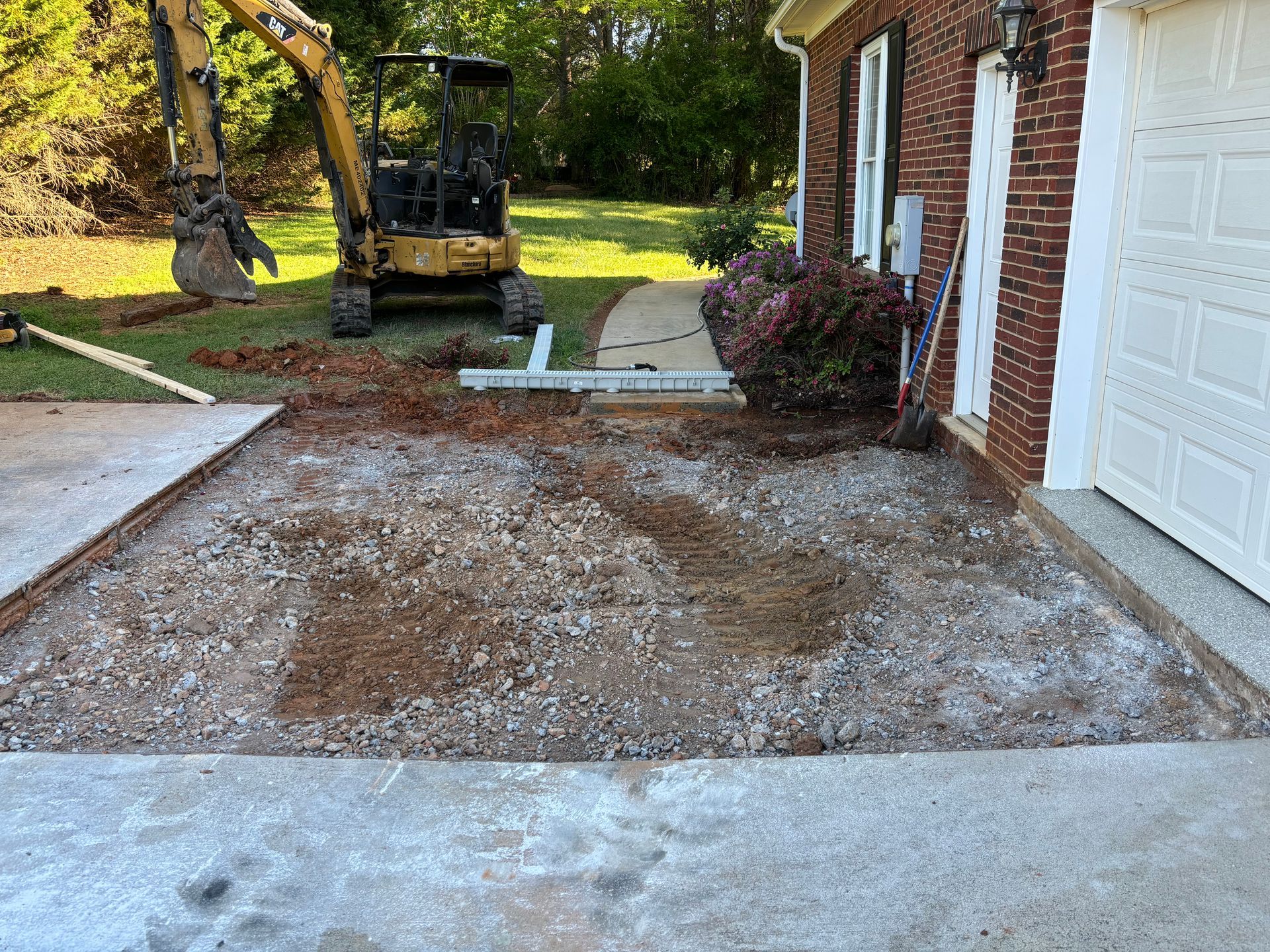 Front yard under renovation beside a brick house, with exposed soil and a compact excavator in the background