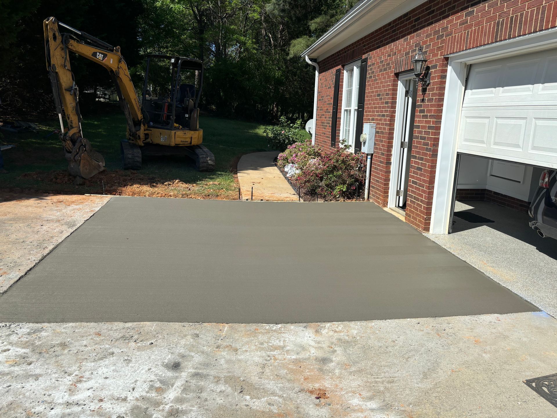 Freshly poured concrete slab beside a brick garage, with an excavator in the driveway.