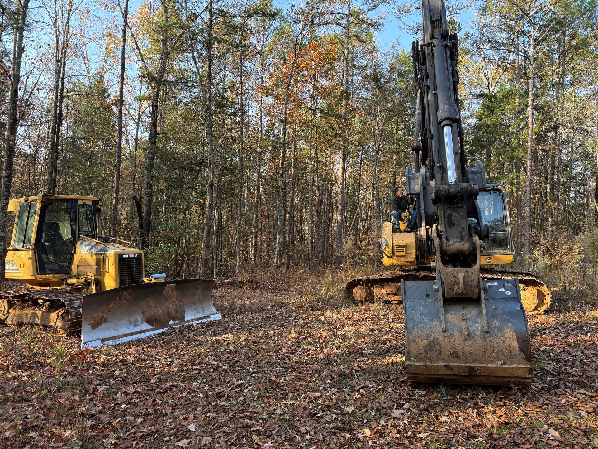 Two yellow excavators clearing leaves in a wooded area in autumn