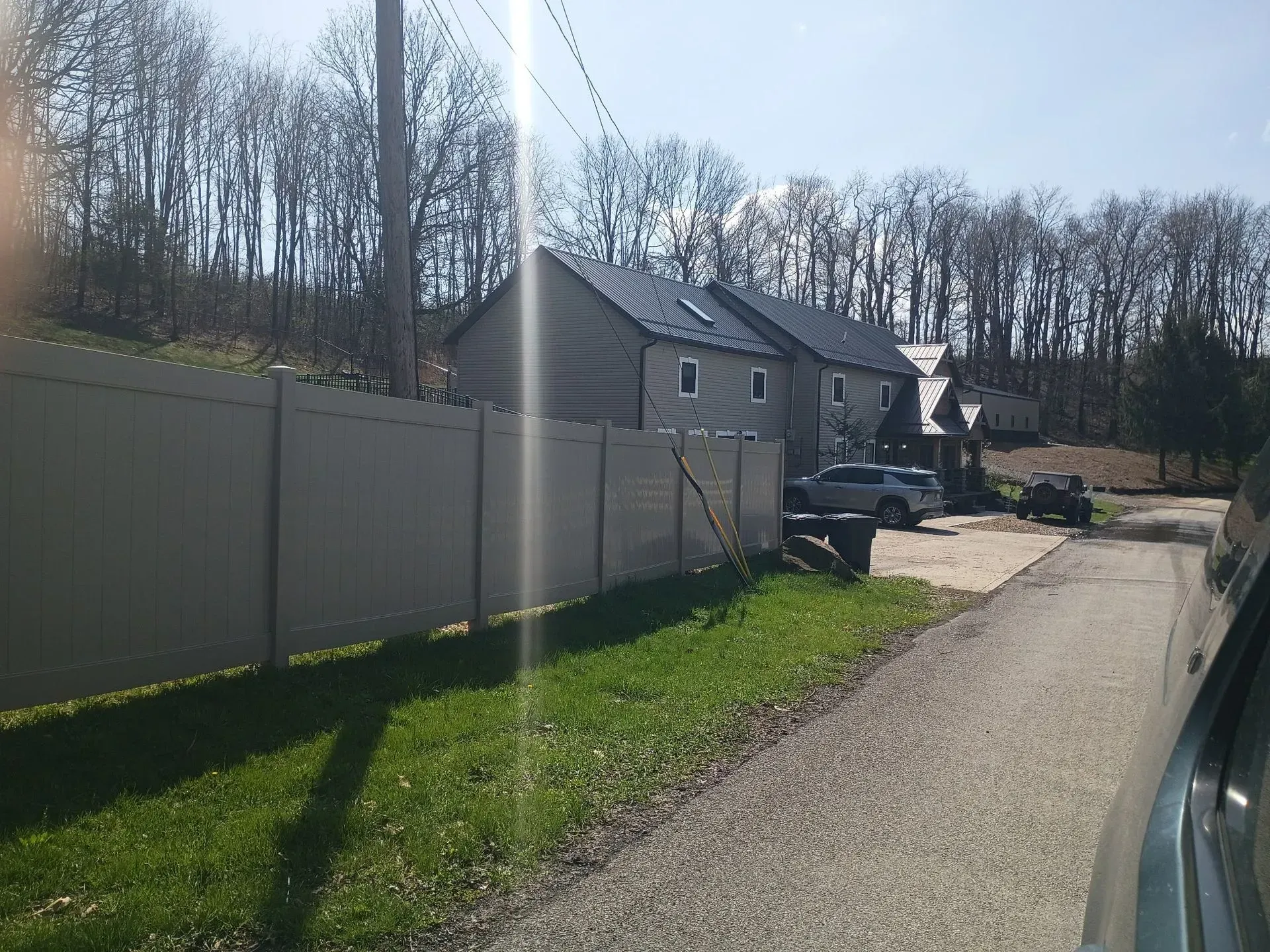 Sunlit driveway beside a white fence, with parked cars and a house in the background