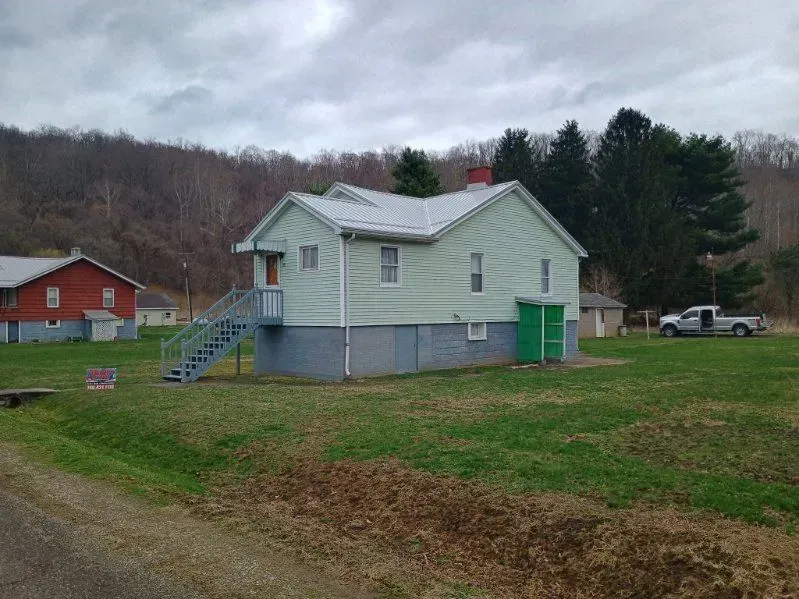 Small pale green house with gray roof on a grassy hillside, with trees and a red barn in the background