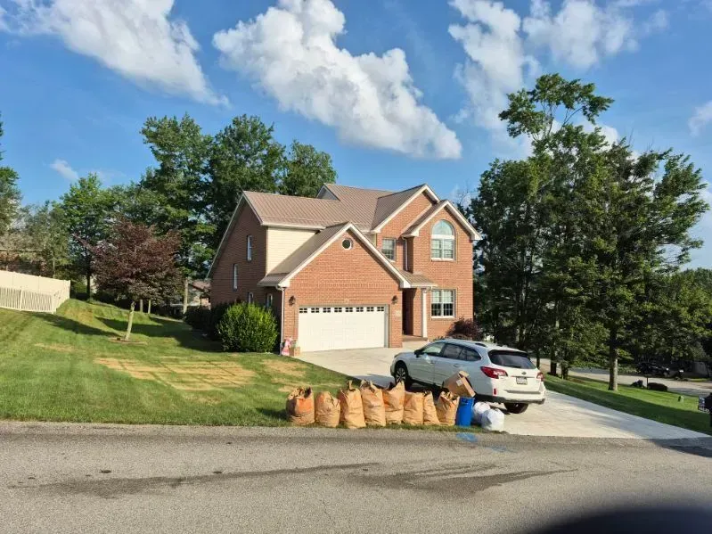 Suburban brick house with white garage, parked SUV, and trash bags at curb on a sunny day