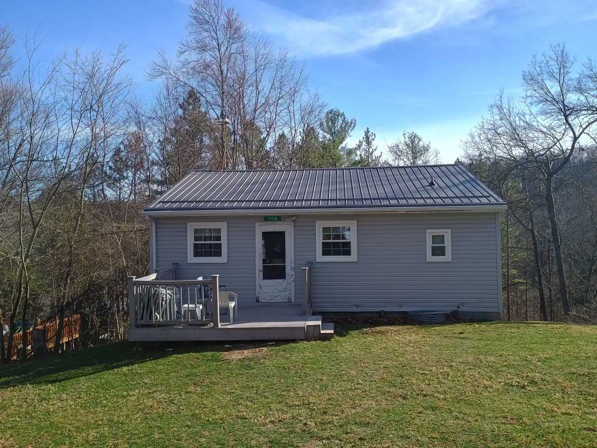 Small gray house with a porch on a grassy hill under a blue sky, surrounded by leafless trees