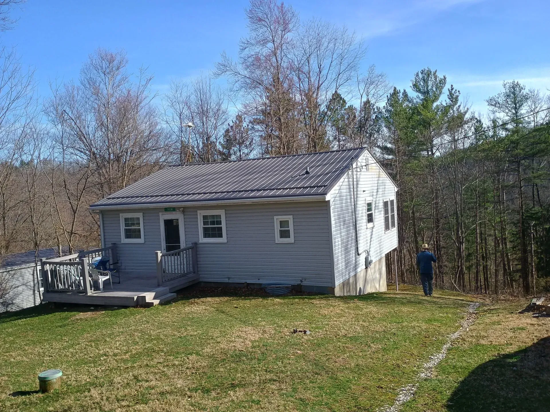 Small gray house with a deck in a grassy yard, with trees and a person standing near the side.