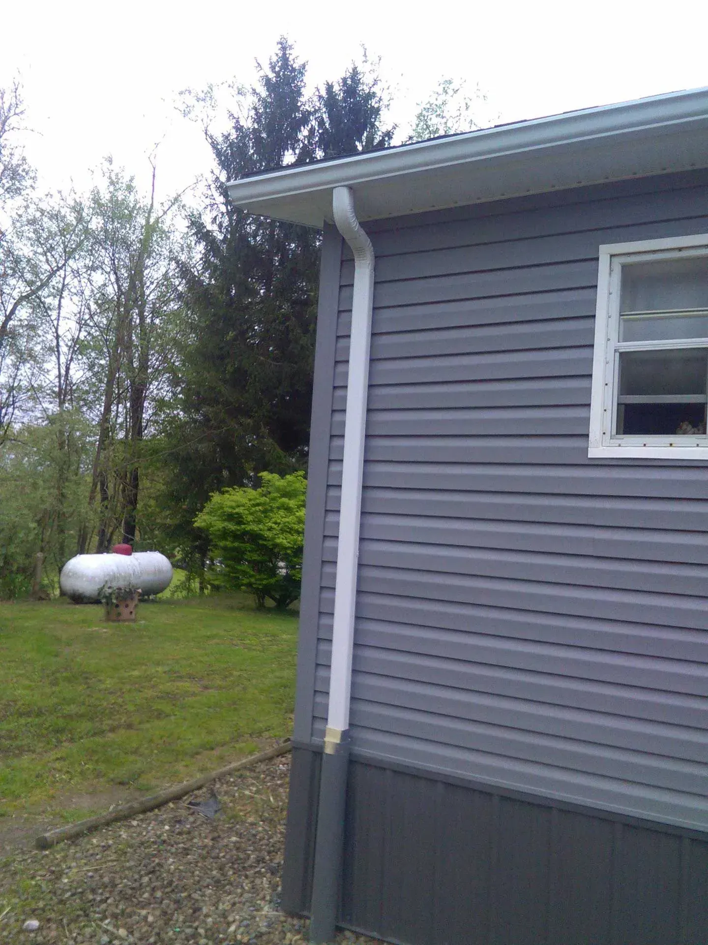 Side of gray house with white trim, small window, gravel path, and a propane tank in a grassy yard.
