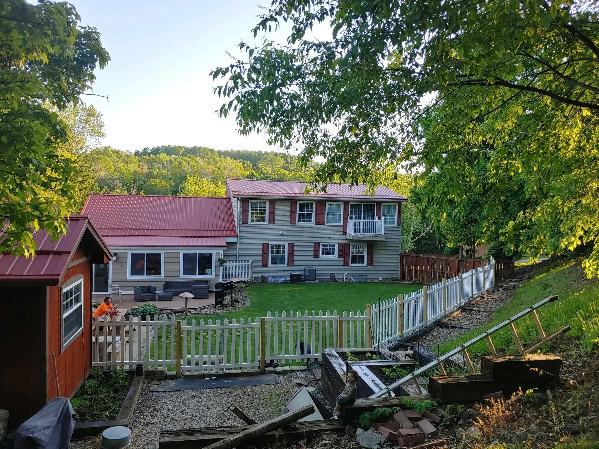 Riverside house with red roof, white fence, and fallen deck railings in a wooded yard
