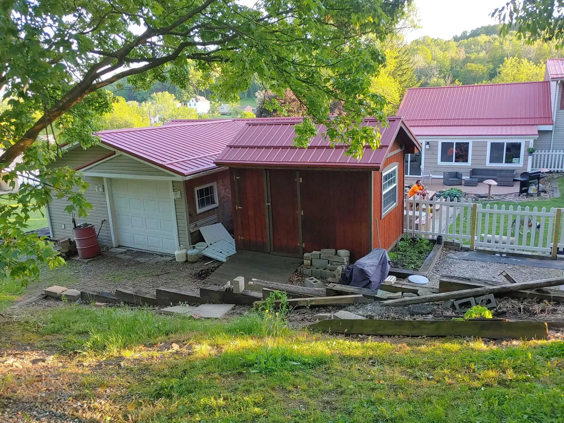 Backyard with a white shed, red-roofed outbuilding, stone wall, and a house in the background.