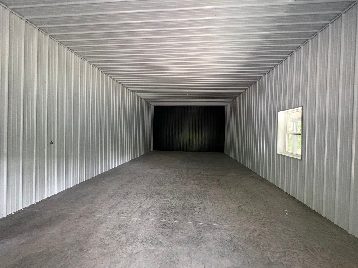 Interior of a long, narrow metal structure, possibly a storage container. Dark floor, corrugated walls, small window.