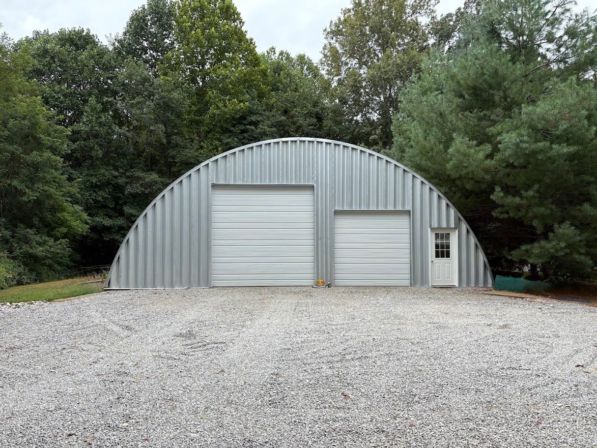 Quonset hut with two garage doors and a small door, set on a gravel driveway, trees in the background.
