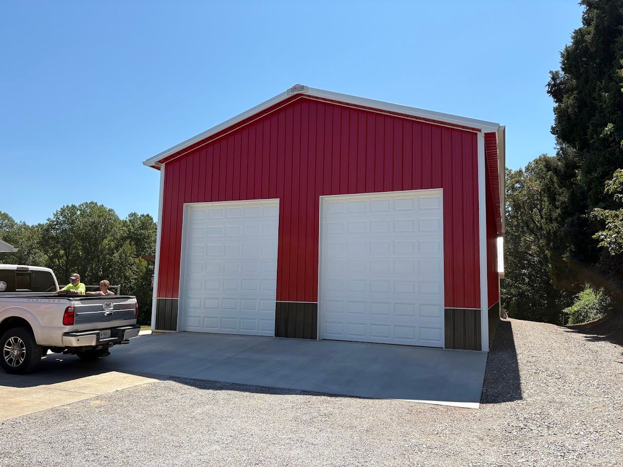 Red metal garage with two white garage doors, a pickup truck, and gravel driveway.