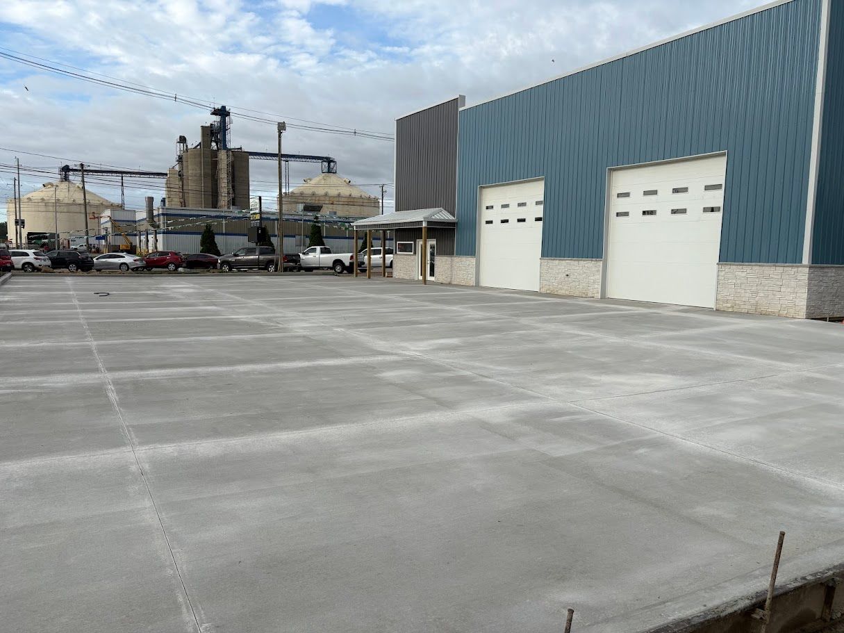 Large, empty concrete lot in front of a blue industrial building with two loading doors, trucks parked nearby.