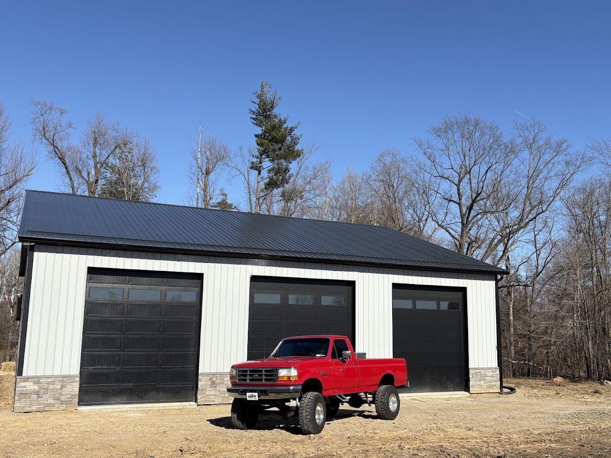 Red pickup truck parked in front of a three-bay garage with black doors and a dark roof under a bright blue sky.