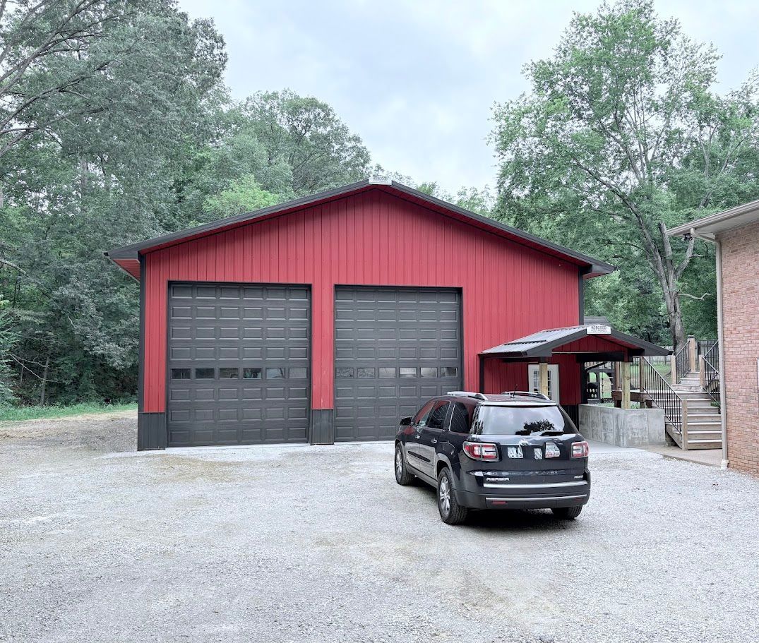 Red garage with two doors, black SUV in front, gravel driveway, trees in the background.