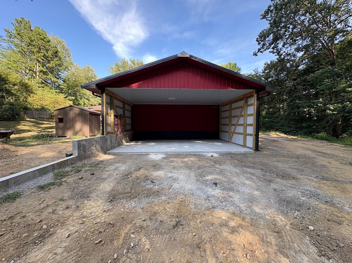 Red and white open-air shed with concrete floor, surrounded by dirt and trees, under a blue sky.