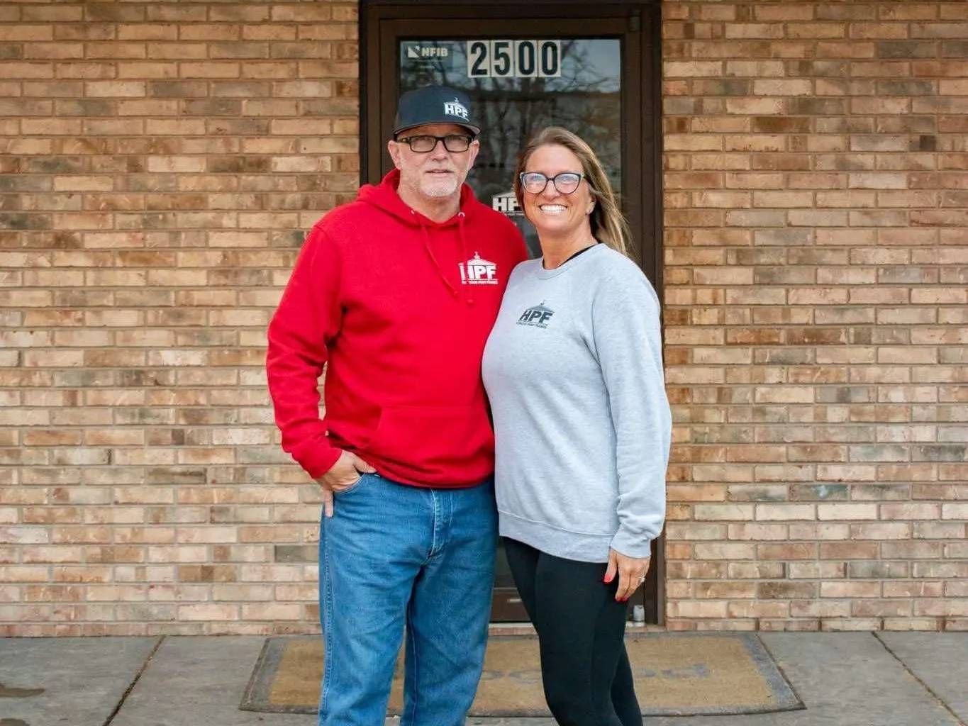 Man and woman in front of a brick building. Man in red hoodie, woman in gray sweatshirt, smiling. Building has a dark door.