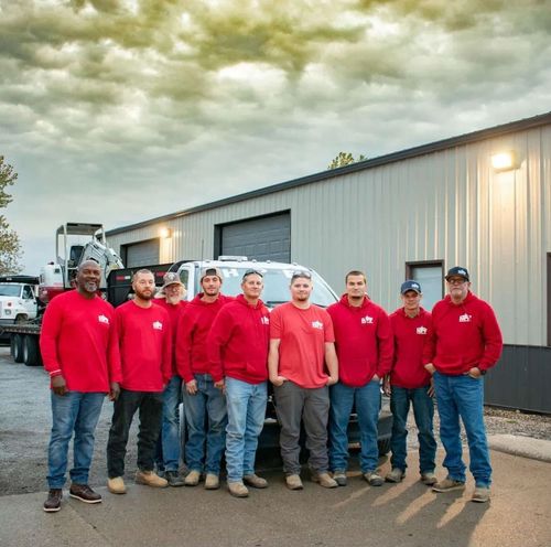 Group of men in red shirts and jeans stand in front of a building and trucks.