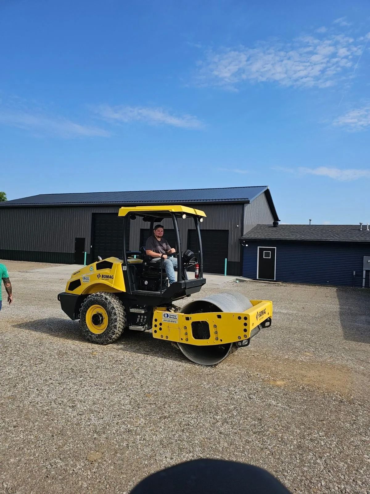 Man driving a yellow road roller on gravel, blue sky and buildings in the background.