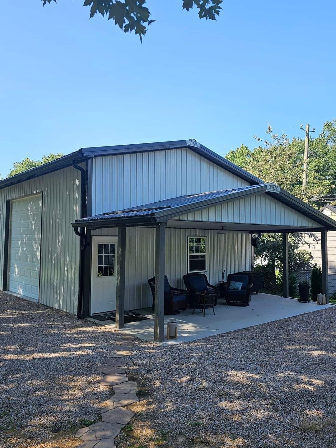 Metal building with a covered patio, gravel path, and trees under a blue sky.