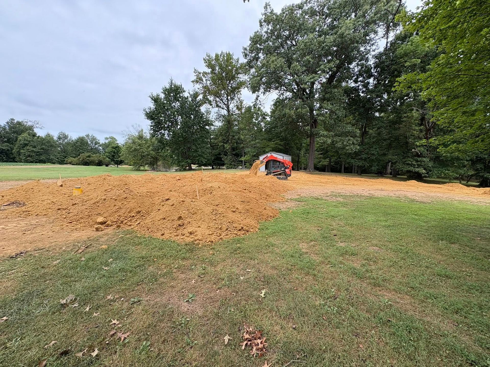Field with upturned dirt patch, small tractor, and trees under a cloudy sky.