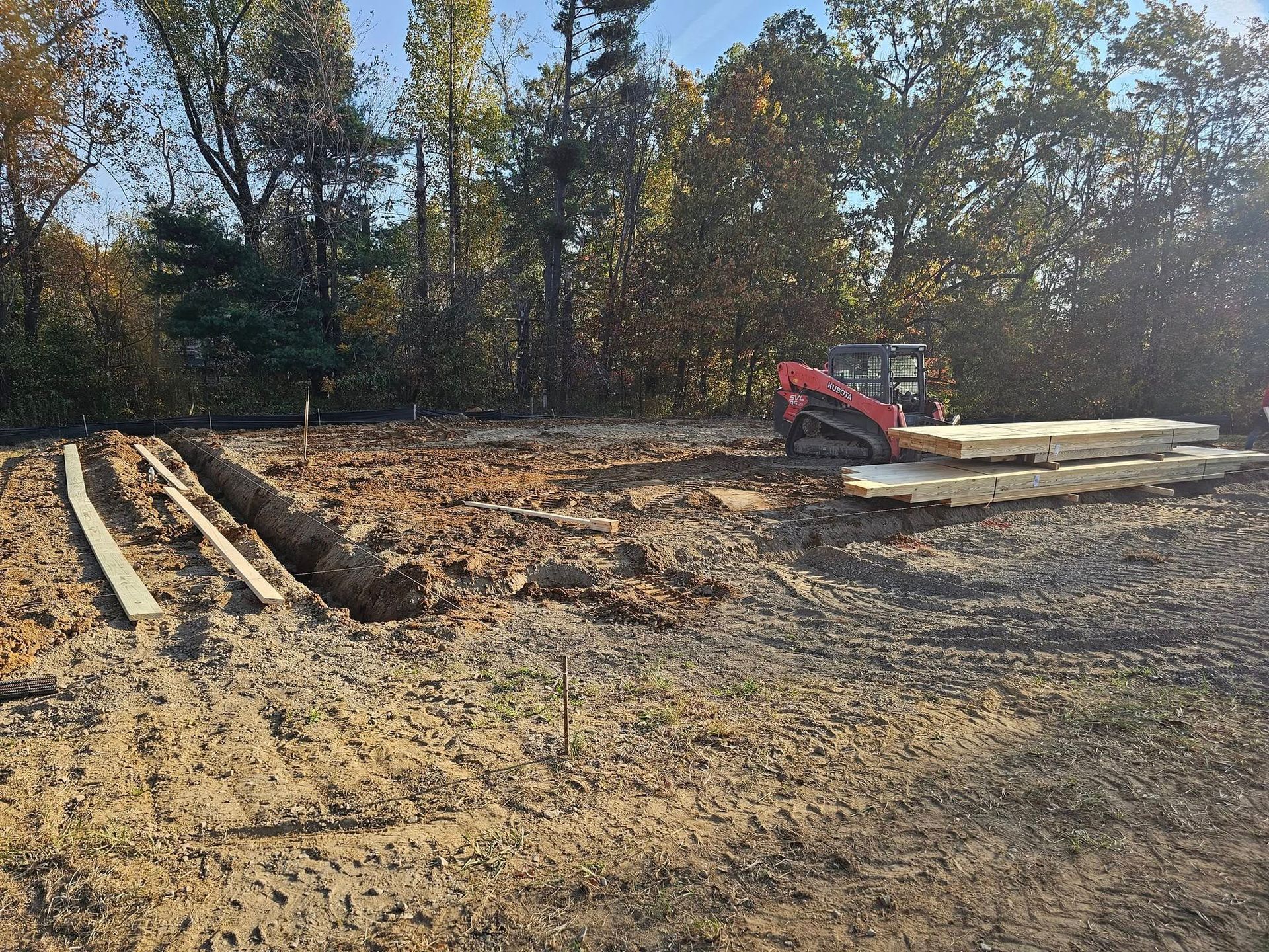 Dirt worksite with small excavator, wooden planks, and trench. Trees in the background under a blue sky.