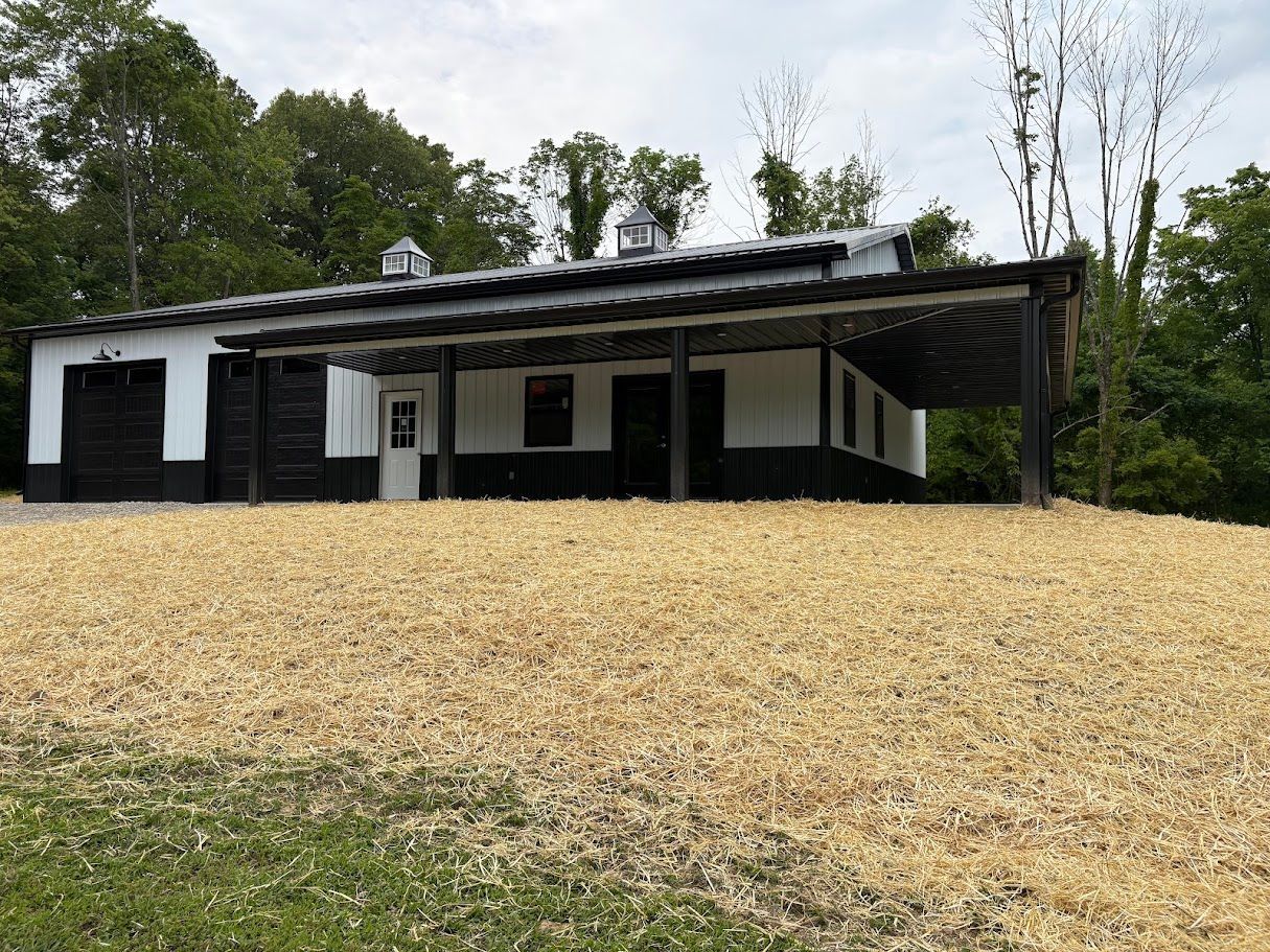 White and black building with a covered porch, set on a grassy hill with trees in the background.