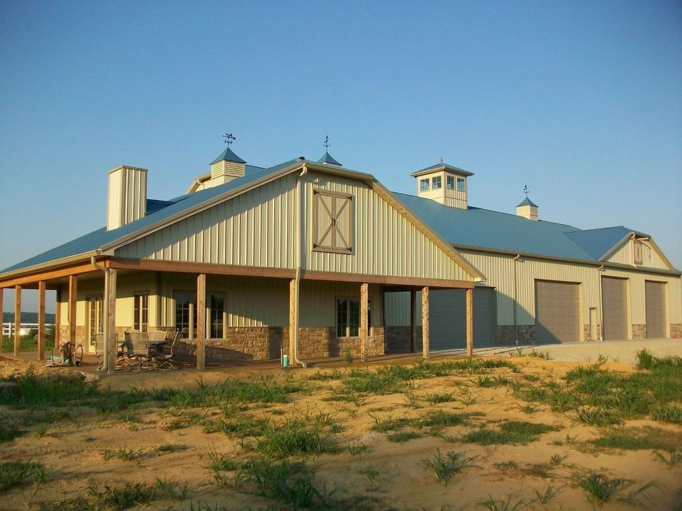 Barn-style building with blue roof, beige siding, stone base, porch, and garage doors, set in a field.