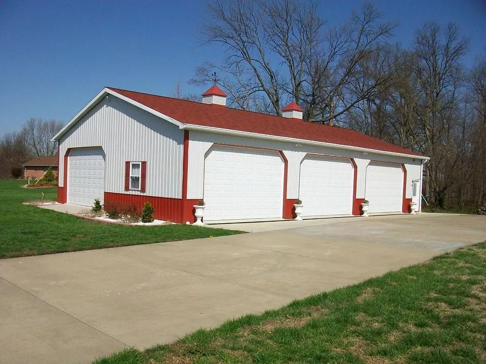 White and red metal garage with four bays, red roof, concrete driveway, green lawn, and trees.