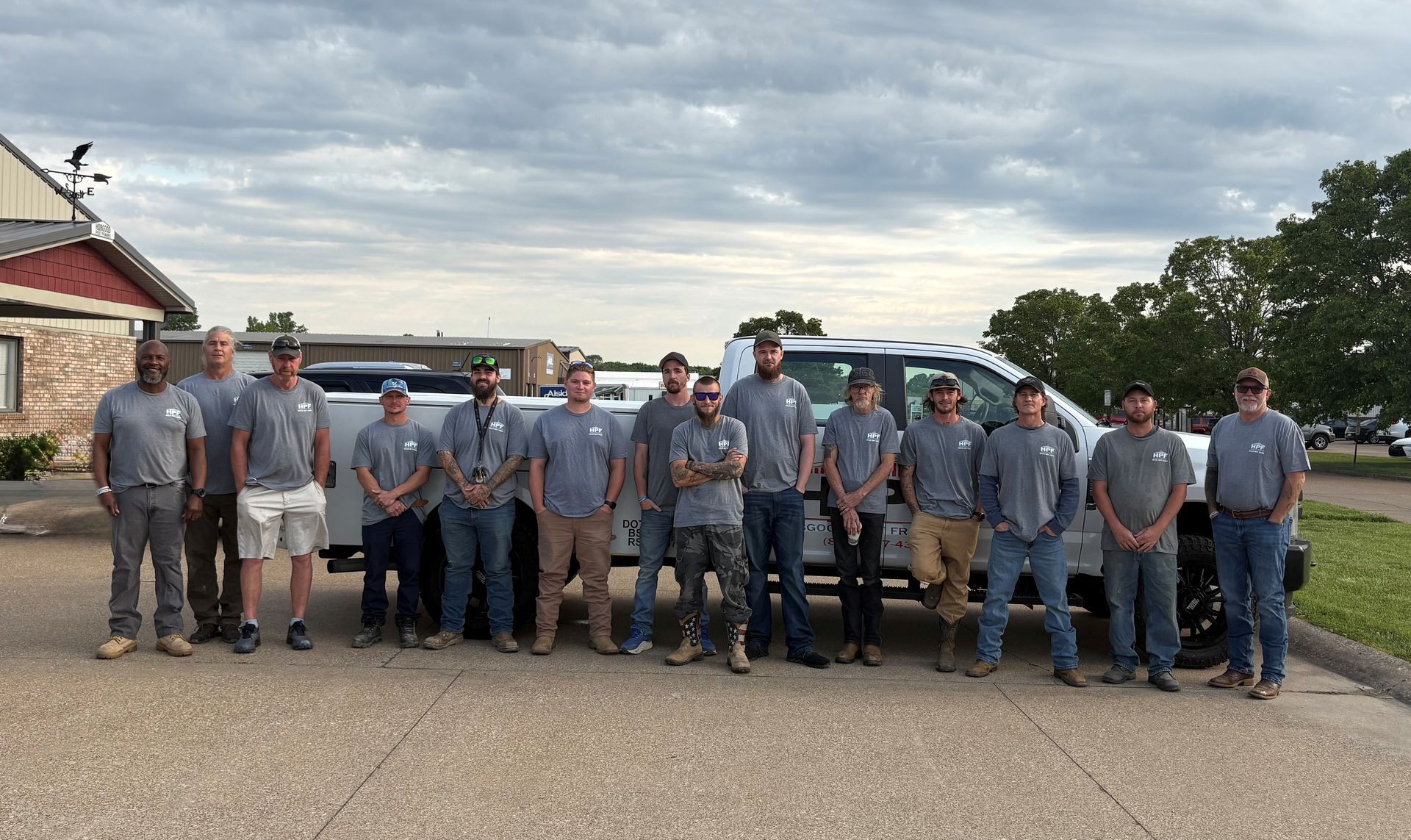 Group of people in gray uniforms standing together outdoors in front of parked trucks and a building