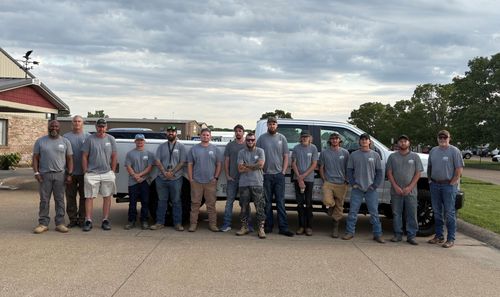 Group of people in gray uniforms standing together outdoors in front of parked trucks and a building