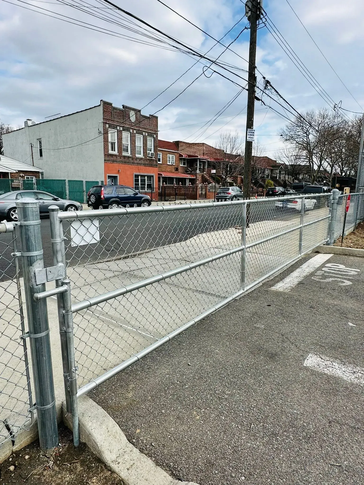 Chain-link fence in front of a parking area. Buildings and power lines in the background.