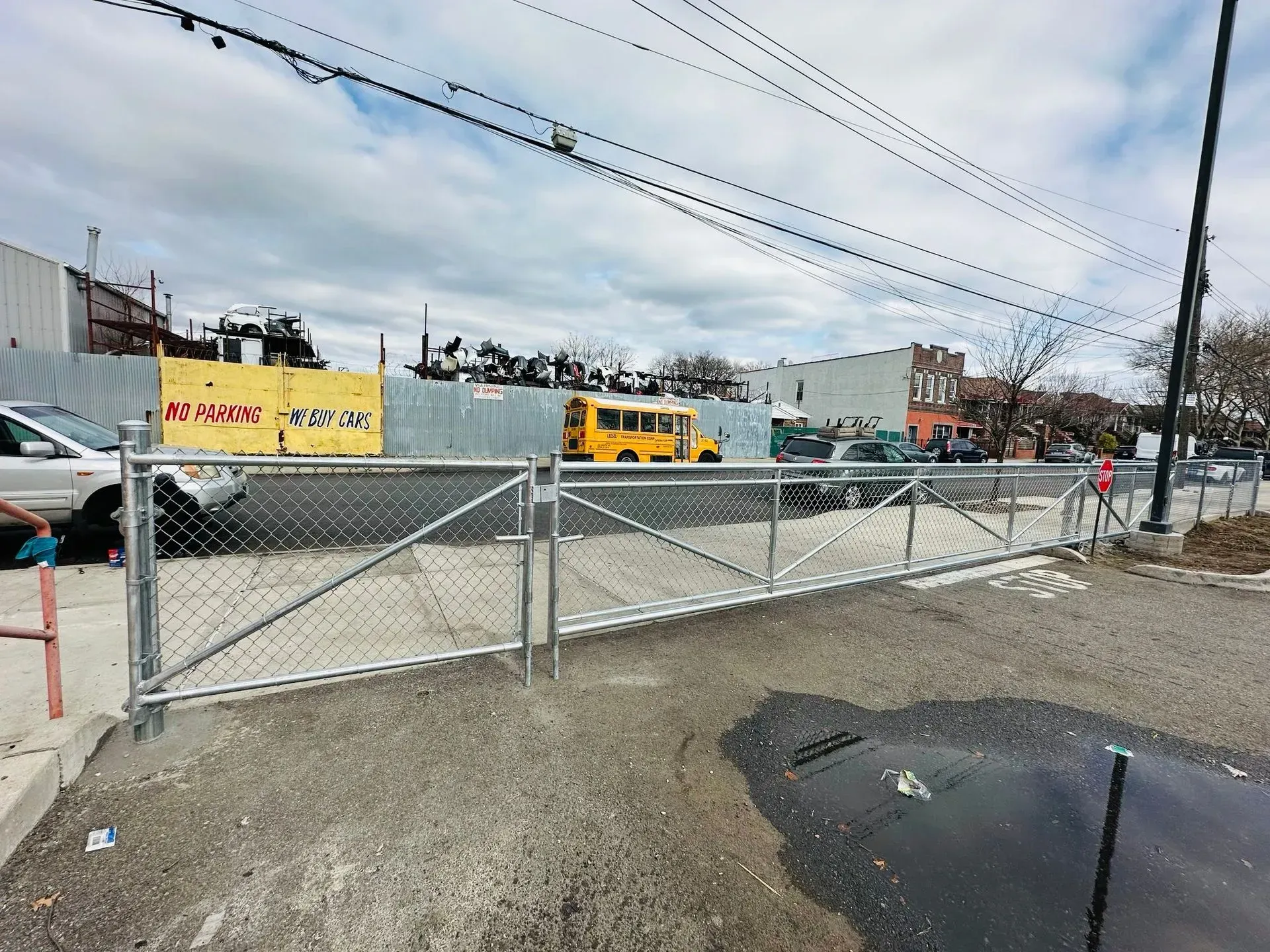 Chain-link fence in front of a gravel lot with a school bus, buildings, and utility wires on an overcast day.