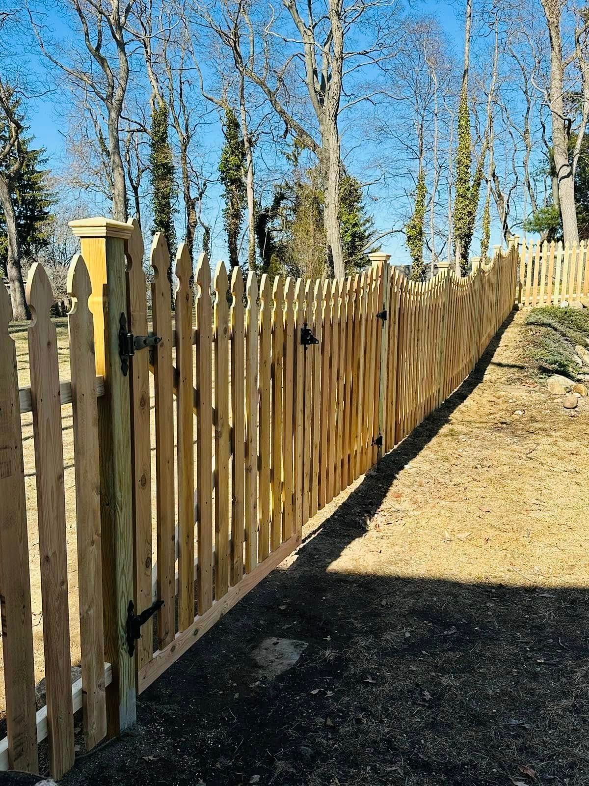 Wooden picket fence with gate, along a yard with trees and blue sky.