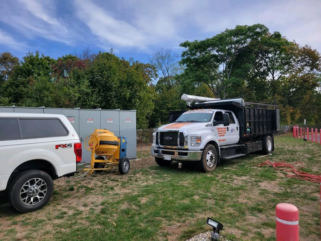White dump truck and Ford SUV parked on grass with construction equipment, trees, and sky.