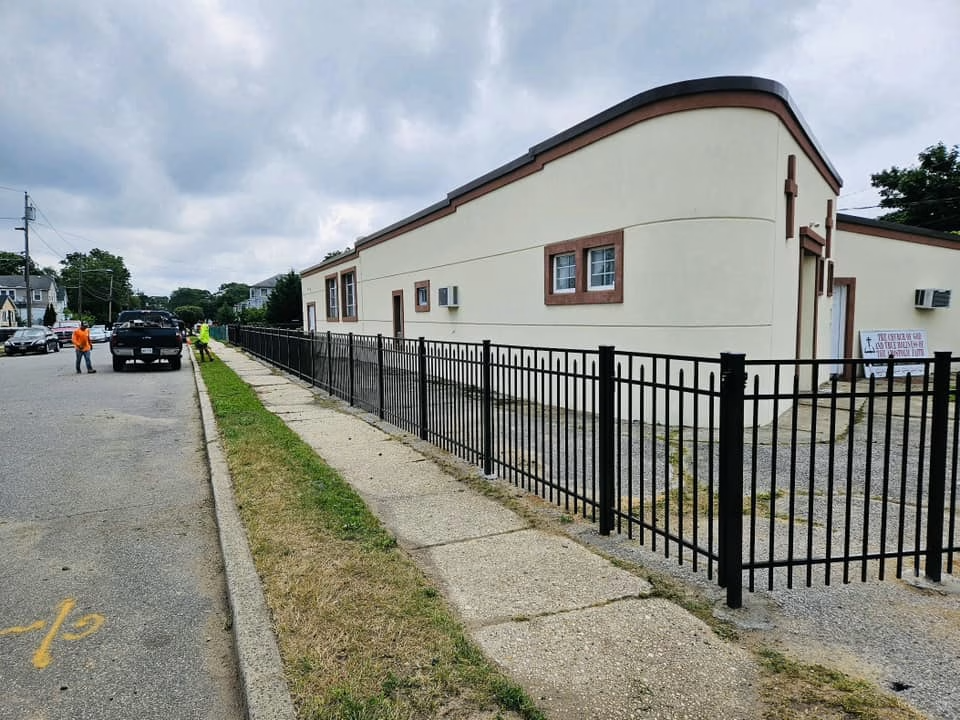 Black metal fence along sidewalk next to tan building with brown trim. Cloudy day, street on left.