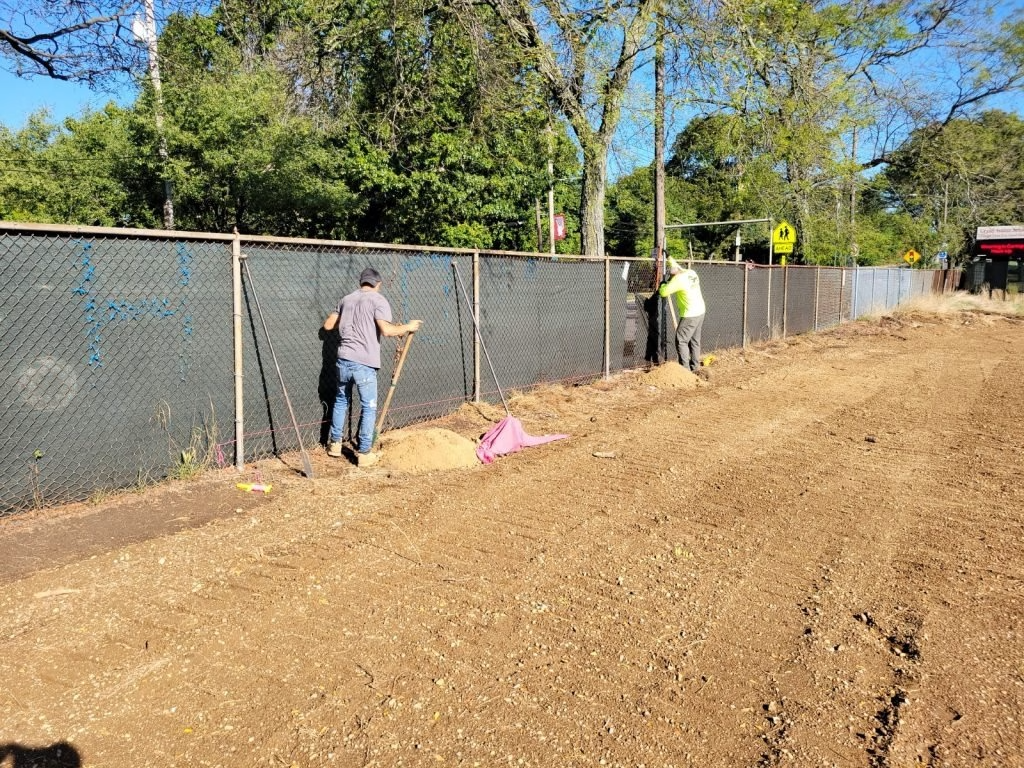 Two workers install a fence along a dirt road, one using a shovel. Green trees and blue sky in background.